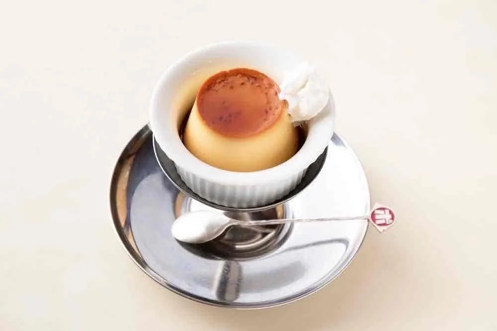 プリンが白い器に盛られ、銀色のソーサーとスプーンの上に置かれています。プリンの上にはカラメルソースと生クリームが添えられています。