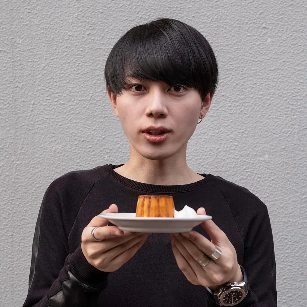 男性が皿に乗ったデザートを持っている写真です。男性は黒い服を着ていて、正面を向いています。デザートは、カラメルソースがかかったプリンのようです。