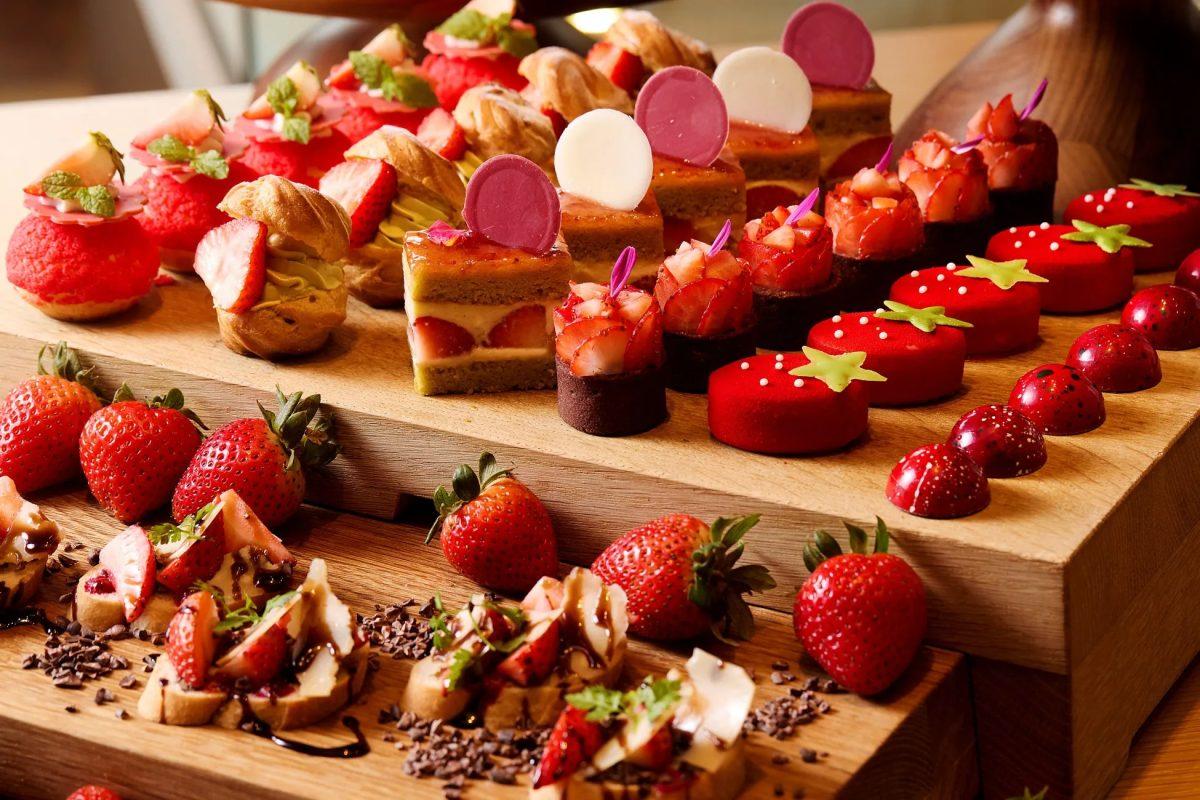 木製の台の上に、いちごをあしらった様々なデザートが並べられた写真。シュークリーム、ケーキ、チョコレートなど、色とりどりのスイーツが美味しそうに配置されている。