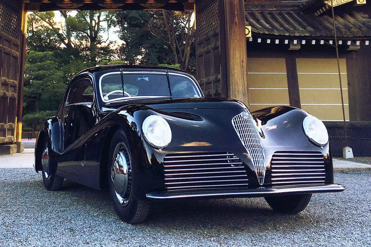 黒いクラシックカーが、日本の伝統的な建築様式である門の前に駐車されている写真です
