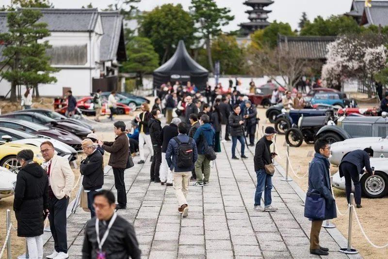 クラシックカーのイベントの様子。多くの人が集まり、様々な種類のクラシックカーが展示されている。背景には日本の寺院のような建物が見える