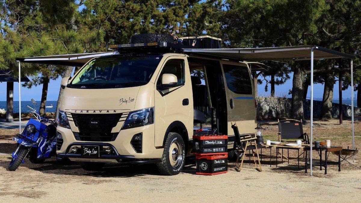 砂浜に停められたカスタムされたキャラバンと、キャンプ用のテーブルや椅子、バイクなどが写った写真です。キャラバンはベージュ色で、屋根には荷物やタイヤが積まれ、サイドにはオーニングが展開されています。キャンプ用品が配置され、アウトドアライフスタイルを想像させる写真です。
